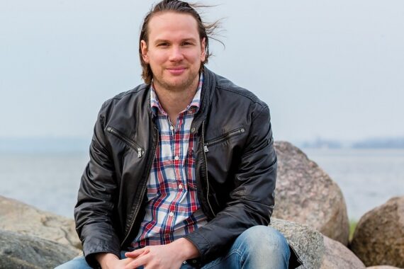 Ein Mann mit langen Haaren sitzt auf einem Felsen am Wasser und trägt eine schwarze Lederjacke, ein kariertes Hemd und eine Jeans. Er lächelt leicht und hat die Hände vor sich verschränkt. Der Himmel und das Wasser im Hintergrund erscheinen bewölkt.