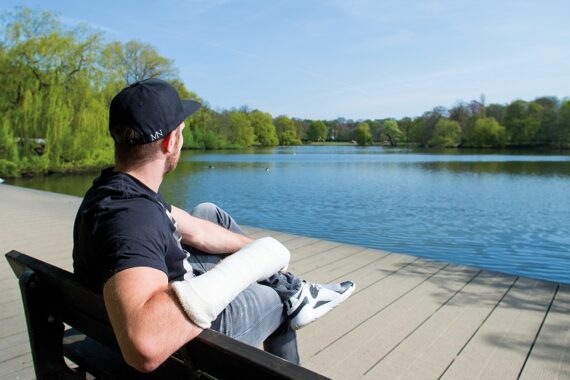 Ein Mann mit einem Gips am rechten Arm sitzt auf einer Bank an einem See und schaut auf das Wasser. Er trägt eine schwarze Mütze, ein schwarzes T-Shirt und eine Jeans. Bäume und Grünflächen umgeben die friedliche Szene am See.