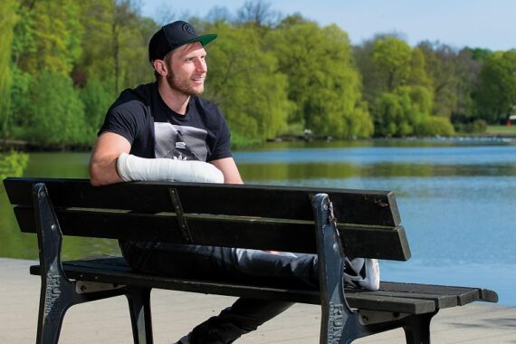 Ein Mann mit einem Gips am linken Arm sitzt auf einer Parkbank an einem See, lächelt und trägt eine schwarze Mütze, ein schwarzes T-Shirt und Jeans. Üppig grüne Bäume umgeben das Wasser an einem sonnigen Tag.