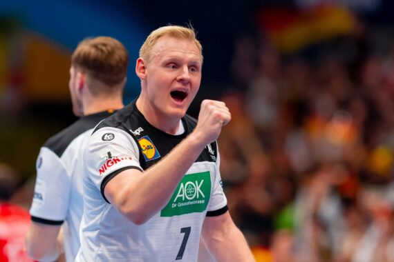 Ein männlicher Handballspieler in einem weiß-schwarzen Trikot mit der Nummer 7 ballt während eines Spiels die Faust und jubelt, während im Hintergrund eine verschwommene Menschenmenge zu sehen ist.
