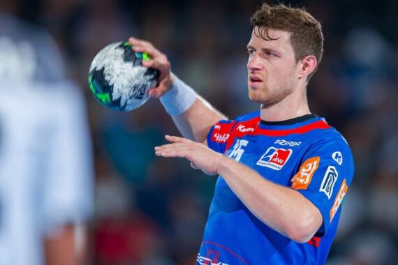 Ein männlicher Handballspieler in einem blau-roten Trikot bereitet sich darauf vor, einen Ball während eines Spiels zu werfen. Er ist konzentriert, hat den rechten Arm erhoben und den Ball in der linken Hand. Der Hintergrund ist unscharf und zeigt einen anderen Spieler.