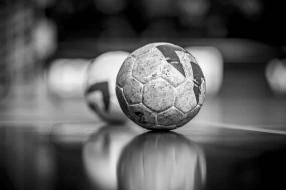 Ein Schwarz-Weiß-Foto in Nahaufnahme eines abgenutzten Handballs, der auf einer glänzenden Hallenfläche liegt, mit einem anderen Handball im Hintergrund, der unscharf abgebildet ist. Die Szene ist weich fokussiert und hebt die Textur des Balls hervor.