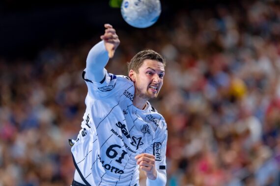 Ein männlicher Handballspieler in einem weißen Trikot wirft in der Mitte des Spiels einen Ball mit voller Wucht. Der Hintergrund ist unscharf und zeigt eine große Menge von Zuschauern.