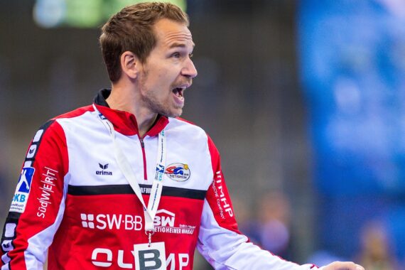 Ein Mann in einer rot-weiß-schwarzen Sportjacke gestikuliert energisch und spricht, als würde er während einer Sportveranstaltung Anweisungen geben. Im Hintergrund ist ein verschwommenes Publikum zu sehen.
