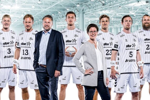 Sieben Personen posieren in einem Innenraum; fünf Männer in passenden weißen Sportuniformen und zwei Personen, ein Mann im Anzug und eine Frau in Geschäftskleidung, stehen davor. Der Hintergrund ist ein großes, hell erleuchtetes Gebäude.