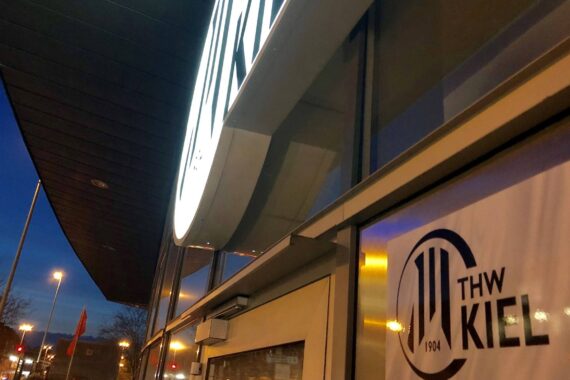 Eingang eines modernen Gebäudes mit einem großen beleuchteten THW-Kiel-Logoschild über der Tür und einem aufgedruckten THW-Kiel-Logo auf der Glasscheibe, fotografiert in der Abenddämmerung bei Straßenbeleuchtung und tiefblauem Himmel.