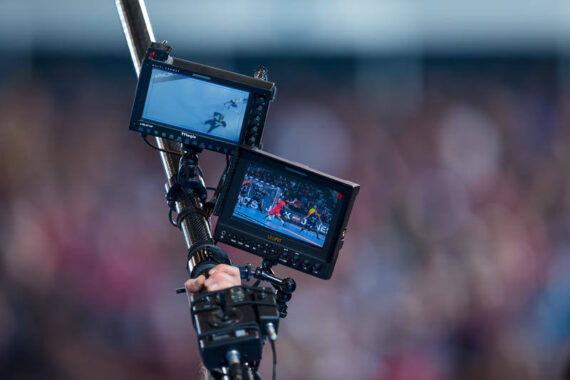 Zwei kleine Monitore auf einer Kameraausrüstung zeigen Live-Aufnahmen eines Sportereignisses, einer zeigt eine Skiszene, der andere ein Stadion mit Sportlern. Der Hintergrund ist mit jubelnden Zuschauern verschwommen.