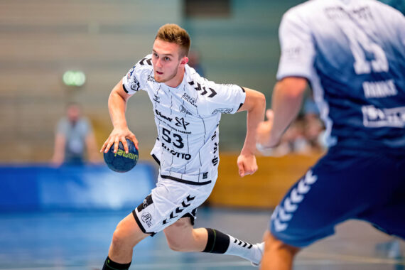 Ein männlicher Handballspieler in einem weißen Trikot sprintet mit dem Ball nach vorne, während ein Spieler in Blau, der teilweise sichtbar ist, verteidigt. Das Geschehen findet in einer Halle auf einem Spielfeld statt, wobei die Zuschauer im Hintergrund verschwimmen.