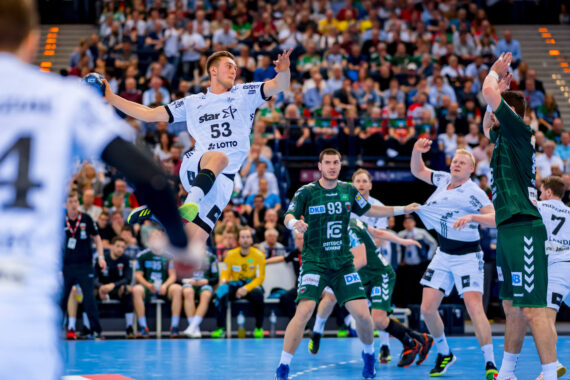 Ein Handballspieler in einem weißen Trikot springt in der Luft, um den Ball zu werfen, während Verteidiger in grünen Trikots und andere Spieler zuschauen, alles auf einem blauen Hallenplatz mit einer jubelnden Menge im Hintergrund.