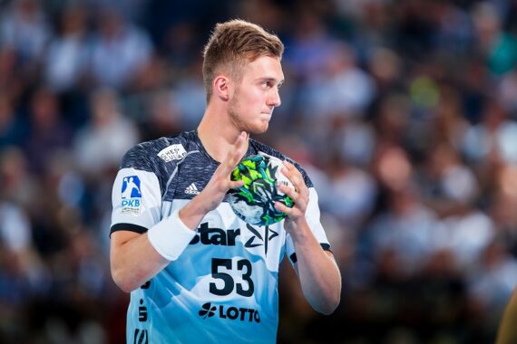 Ein männlicher Handballspieler in einem hellblauen Trikot mit der Nummer 53 hält einen Handball, konzentriert während eines Spiels. Der Hintergrund zeigt eine unscharfe Menge und eine Halle.