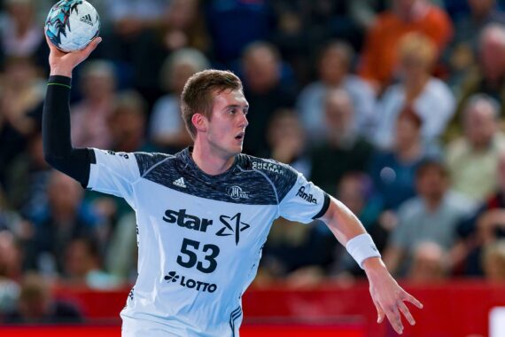 Ein männlicher Handballspieler in einem weiß-marineblauen Trikot mit der Nummer 53 bereitet sich darauf vor, den Ball während eines Spiels zu werfen. Im Hintergrund ist eine unscharfe Menschenmenge zu sehen.