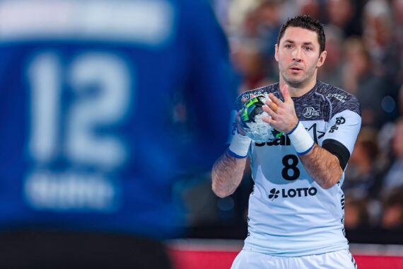 Ein männlicher Handballspieler in einem weißen Trikot mit der Nummer 8 hält den Ball und bereitet sich auf das Spiel vor, während ein anderer Spieler in einem blauen Trikot mit der Nummer 12 im Vordergrund steht, leicht unscharf.