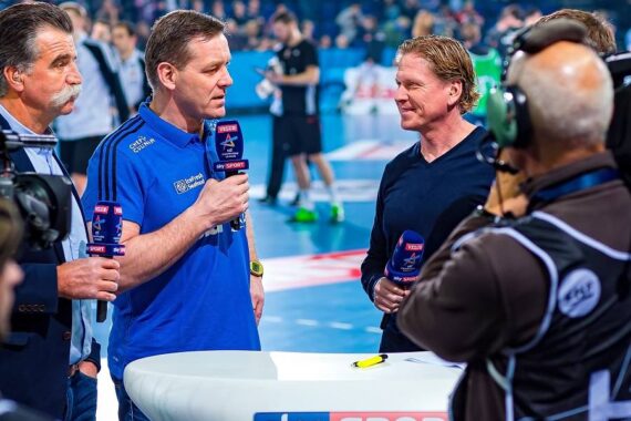 Vier Männer stehen um einen Tisch mit dem Logo von Sky Sport und halten Mikrofone während eines im Fernsehen übertragenen Sportinterviews. Ein Kameramann filmt das Gespräch in einer hell erleuchteten Halle.