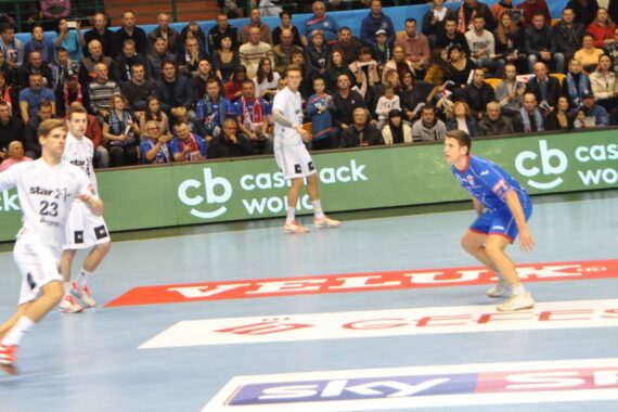 Ein Handballspiel ist im Gange. Ein Spieler in weißer Uniform bereitet sich auf den Wurf vor, während ein Spieler in Blau den Ball verteidigt. Die Zuschauer füllen die Tribünen im Hintergrund, und um das Spielfeld herum sind verschiedene Werbungen zu sehen.