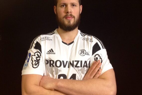 Ein männlicher Sportler mit Bart steht mit verschränkten Armen vor einem schwarzen Hintergrund und trägt ein weiß-schwarzes Sporttrikot mit der Aufschrift PROVINZIAL und verschiedenen Logos darauf.