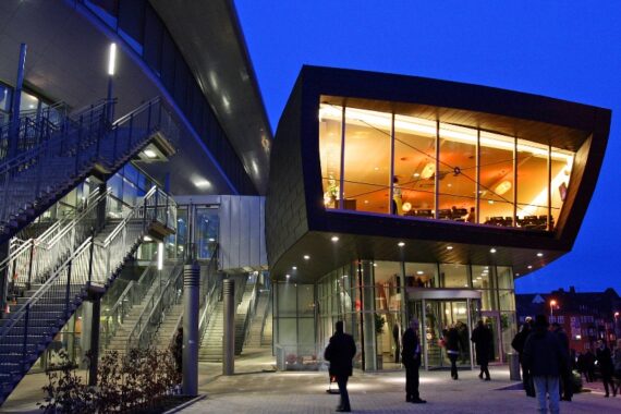 Ein modernes Gebäude mit Glaswänden und einem beleuchteten Obergeschoss in der Abenddämmerung. Menschen stehen draußen in der Nähe des Eingangs und entlang einer Metalltreppe, während der Himmel tiefblau ist.