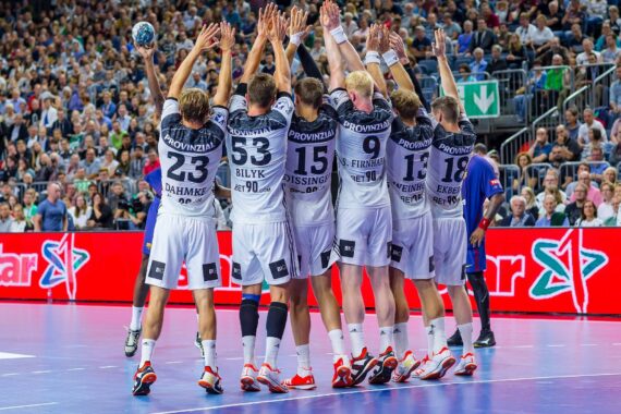Eine Handballmannschaft in weißen Trikots steht in einer Reihe mit erhobenen Armen und verteidigt sich gegen einen Gegner in Blau während eines Spiels in einer überfüllten Halle. Die Zuschauer füllen die Tribünen im Hintergrund.