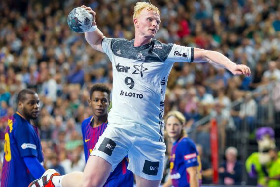 Ein männlicher Handballspieler in einem weißen Trikot springt mit einem Ball in der rechten Hand durch die Luft und bereitet sich auf den Wurf vor, während seine Gegner in blauen und roten Trikots zusehen. Im Hintergrund ist eine unscharfe Zuschauermenge zu sehen.