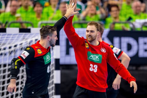 Zwei Handballer in schwarz-roten Trikots feiern mit einem High Five vor einem Tornetz, während im Hintergrund Menschen in grünen Hemden von der Tribüne aus zusehen.
