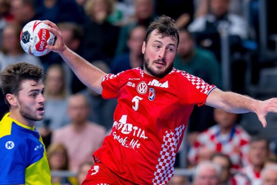 Ein männlicher Handballspieler in einem rot-weißen kroatischen Trikot springt, um den Ball zu werfen, während ein Spieler in einem gelb-blauen Trikot zusieht. Die Zuschauer sind im Hintergrund verschwommen.