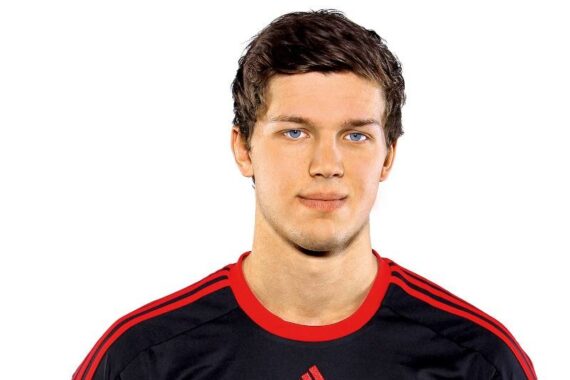 Ein junger Mann mit kurzen braunen Haaren, der ein schwarzes Adidas-Sportshirt mit roten Akzenten trägt, steht vor einem schlichten weißen Hintergrund und blickt mit neutralem Blick in die Kamera.
