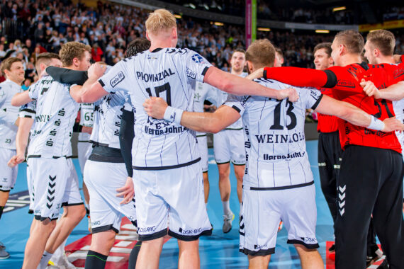Eine Gruppe männlicher Handballspieler in weißen und schwarzen Trikots steht im Kreis und legt die Arme umeinander, um vor einer jubelnden Menge auf einem Hallenplatz zu feiern.