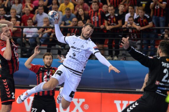 Ein Handballspieler in einem weißen Trikot springt, um den Ball in Richtung Tor zu schießen, während der Torwart in Schwarz sich darauf vorbereitet, den Ball abzuwehren. Die Spieler der gegnerischen Mannschaft in roten und schwarzen Streifen schauen vom Spielfeld aus zu, während im Hintergrund eine Menschenmenge steht.