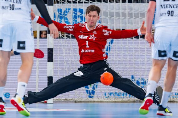 Ein Handballtorwart in einem roten Trikot führt einen Spagat aus, um einen orangefarbenen Ball zu blocken, während zwei gegnerische Spieler in weißen Trikots auf das Tor zugehen. Das Netz und das blaue Spielfeld sind im Hintergrund zu sehen.