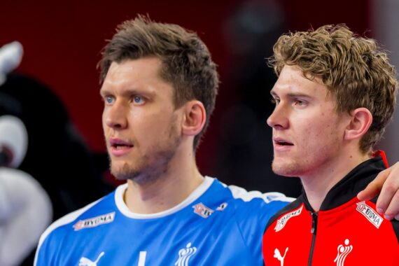 Zwei männliche Sportler stehen nebeneinander, der eine in einem blauen, der andere in einem roten Trikot. Beide blicken konzentriert nach vorne, die Arme um die Schultern des jeweils anderen. Im Hintergrund ist ein verschwommenes Maskottchen zu sehen.