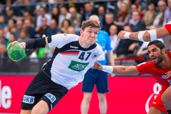 Ein männlicher Handballspieler in einem weiß-schwarzen Trikot, der einen grünen Ball hält, bereitet sich auf den Wurf vor, während er von einem Gegner in einem roten Trikot während eines Hallenspiels verteidigt wird. Im unscharfen Hintergrund sind Zuschauer zu sehen.