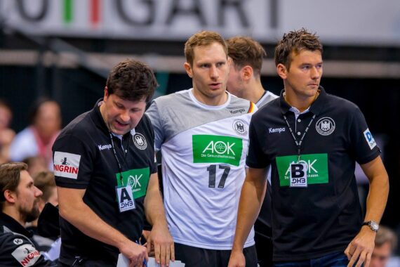 Drei Männer, wahrscheinlich Mitglieder einer Sportmannschaft oder eines Trainerstabs, stehen zusammen und tragen schwarze und weiße Hemden mit Sponsorenlogos. Der Mann in der Mitte trägt ein Trikot mit der Nummer 17. Indoor-Arena, unscharfe Menschenmenge im Hintergrund.