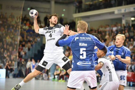 Ein Handballspieler in einem weißen Trikot springt zum Schuss, während Spieler in blauen Trikots versuchen, ihn während eines Hallenspiels zu blockieren, mit einer jubelnden Menge im Hintergrund.