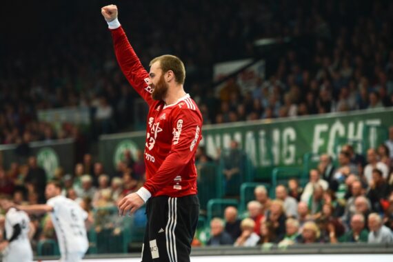 Ein männlicher Handballspieler in einem roten Trikot hebt jubelnd die Faust auf dem Spielfeld, während im Hintergrund eine verschwommene Menschenmenge zusieht.