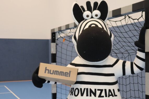 Eine Person in einem Zebra-Maskottchenkostüm mit der Aufschrift PROVINZIAL auf der Brust steht in einer Sporthalle vor einem Handballtor und hält eine braune Schachtel mit der Aufschrift hummel in der Hand.