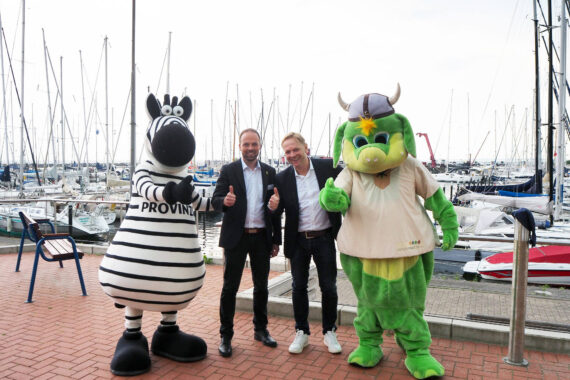 Zwei Männer in Anzügen stehen zwischen einem Zebramaskottchen und einem grünen Drachenmaskottchen, die alle lächeln und Daumen nach oben zeigen, während im Hintergrund ein Yachthafen voller Segelboote zu sehen ist.
