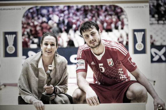 Eine Frau, die einen Pokal in der Hand hält, sitzt neben einem männlichen Sportler in einem roten Trikot mit der Nummer 5. Beide lächeln, im Hintergrund sind eine unscharfe Menschenmenge und Sportbanner zu sehen.
