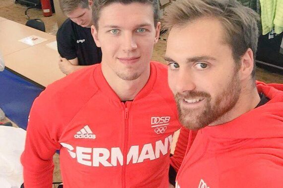 Zwei Männer in roten Jacken mit DEUTSCHLAND- und Sportlogos lächeln für ein Selfie in einer Halle. Ein Mann blickt in die Kamera, während der andere etwas hinter ihm steht.