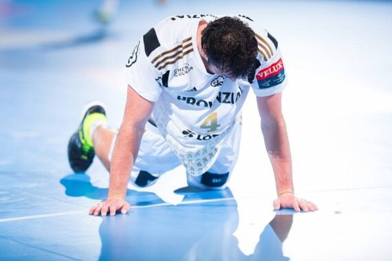 Ein männlicher Handballspieler in einem weißen Trikot mit der Nummer 4 kniet auf dem Spielfeld, stützt sich mit beiden Händen auf dem Boden ab und wirkt während eines Spiels erschöpft oder verletzt.