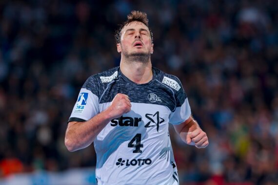 Ein männlicher Handballspieler, der ein weißes und dunkles Trikot mit der Nummer 4 trägt, ballt die Fäuste und blickt nach oben, wobei er während eines Spiels emotional wirkt, mit einer verschwommenen Menschenmenge im Hintergrund.