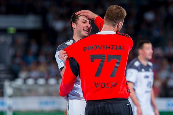 Ein Handballspieler in einem roten Torwarttrikot mit der Aufschrift PROVINZIAL 77 WOLF tätschelt während eines Spiels spielerisch den Kopf eines lächelnden gegnerischen Spielers, während im Hintergrund unscharfe Zuschauer zu sehen sind.
