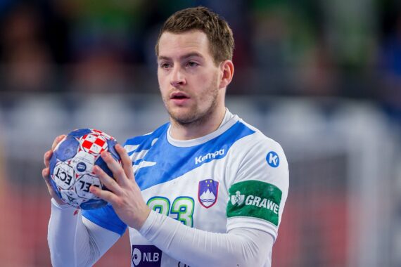 Ein männlicher Handballspieler in einem weiß-blauen Trikot, der mit beiden Händen einen Ball hält, bereitet sich auf den Wurf während eines Spiels vor. Er trägt eine grüne Kapitänsbinde, und der Hintergrund ist mit Zuschauern verschwommen.