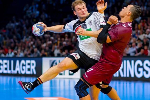 Zwei männliche Handballspieler sind auf dem Spielfeld in Aktion; der Spieler in Weiß springt und hält den Ball, während der Spieler in Kastanienbraun versucht, ihn zu blocken. Im Hintergrund sieht eine Menschenmenge zu.