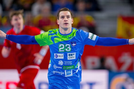 Ein männlicher Handballspieler in einem blau-grünen Trikot mit der Nummer 23 jubelt mit ausgestreckten Armen. Ein anderer Spieler in einem roten Trikot ist unscharf im Hintergrund zu erkennen, und die Zuschauer füllen die Tribüne.