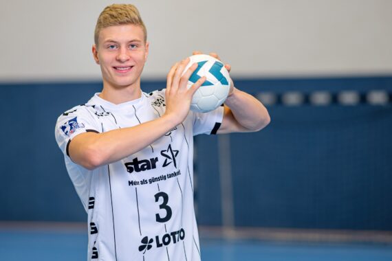 Ein junger männlicher Handballspieler in einem weiß-schwarzen Trikot mit der Nummer 3 lächelt, während er einen Handball mit beiden Händen auf einem Hallenplatz hält. Im Hintergrund ist ein Tornetz zu sehen.