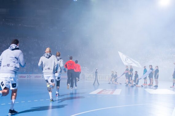Handballspieler in grauen Kapuzenpullovern laufen auf ein verrauchtes Hallenfeld, wo Fans, Schiedsrichter und andere Spieler warten. Helle Lichter und eine jubelnde Menge füllen die Arena im Hintergrund.