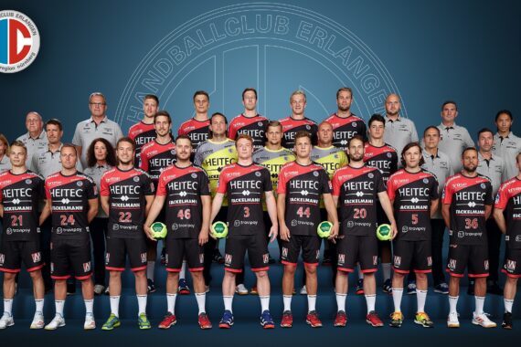 Eine Profi-Handballmannschaft stellt sich für ein Gruppenfoto auf. Vorne stehen die Spieler in schwarzen Trikots mit Handbällen, dahinter die Trainer und Betreuer in grauen Hemden vor einem blauen Hintergrund mit Vereinslogo.