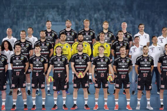 Eine Männerhandballmannschaft in schwarzen Trikots posiert für ein Gruppenfoto mit Trainern und Betreuern, die in Reihen vor einem blauen Hintergrund stehen. Vier Spieler im Vordergrund halten rote und gelbe Bälle.