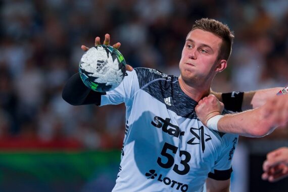 Ein männlicher Handballspieler in einem weißen Trikot mit der Nummer 53 hält einen Ball, während er während eines Spiels von einem gegnerischen Arm um den Hals verteidigt wird. Der Hintergrund ist mit Zuschauern unscharf.