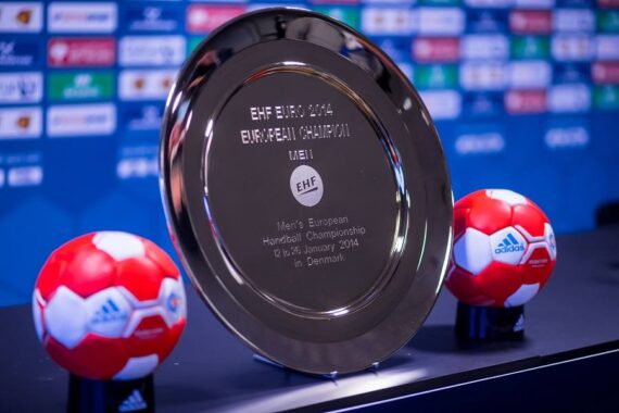 Eine silberne Trophäe mit der Aufschrift EHF EURO 2014 EUROPEAN CHAMPION ist zwischen zwei rot-weißen Adidas-Handbällen zu sehen, mit einem unscharfen Sponsorenhintergrund im Hintergrund.