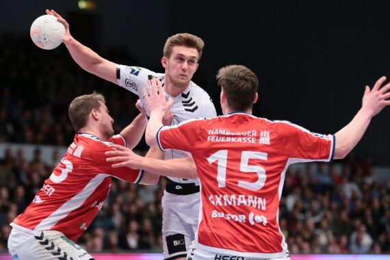 Ein Handballspieler in einem weißen Trikot bereitet sich auf den Wurf vor, während zwei Verteidiger in roten Trikots mit erhobenen Armen ihn während eines intensiven Spiels abwehren, während die Zuschauer im Hintergrund zuschauen.